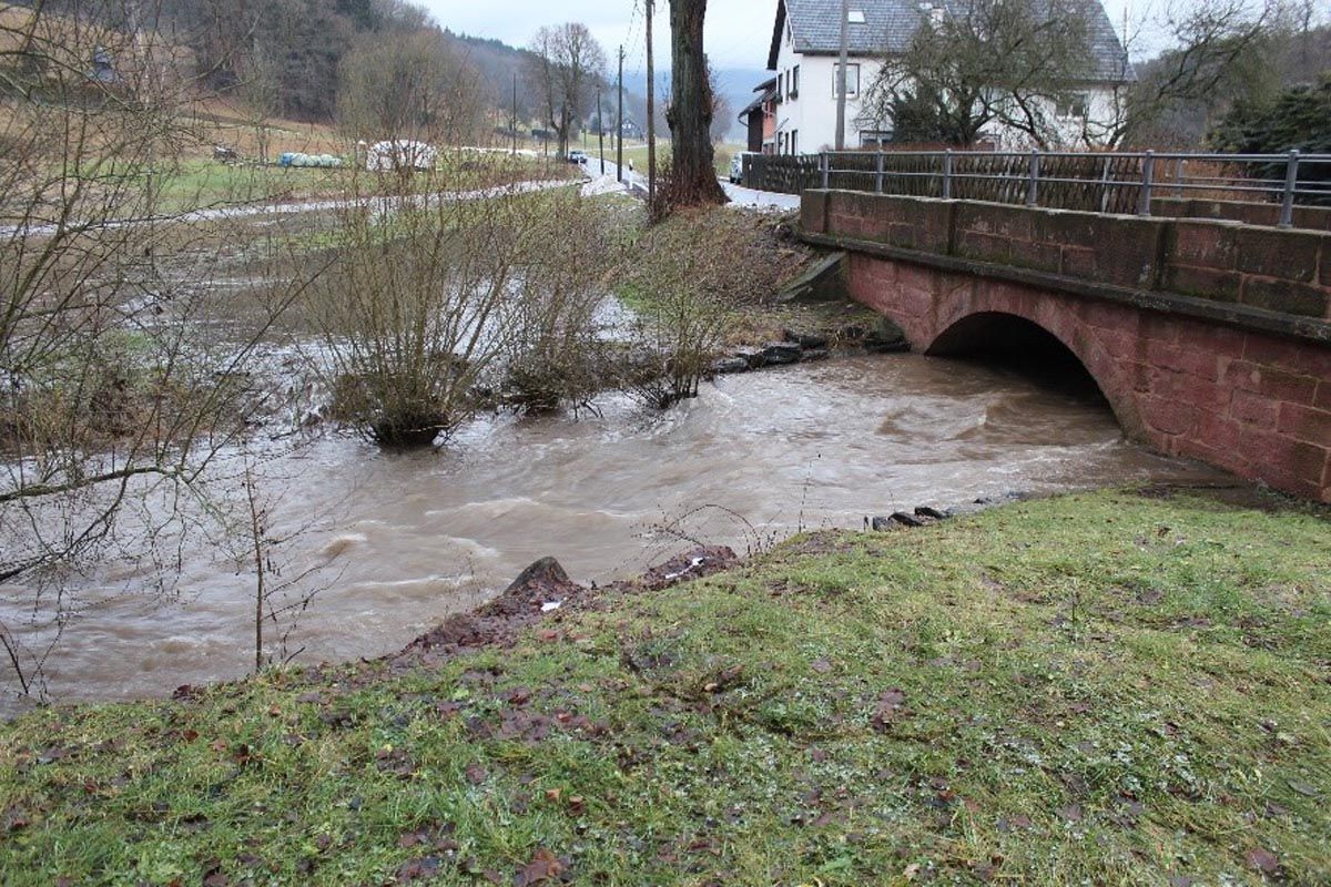 überschwemmte Wiese an steinerner Flussbrücke mit Haus und Straße im Hintergrund