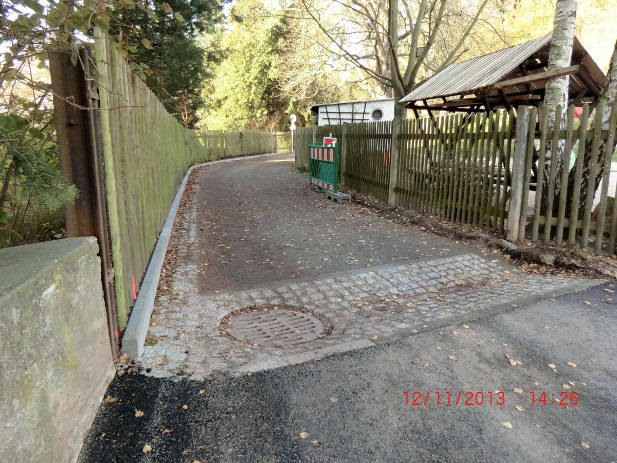 Straße mit Granitpflastermulde und Einlaufgulli; Holzzaun links und rechts; Rehraufe hinter dem rechten Holzzaun